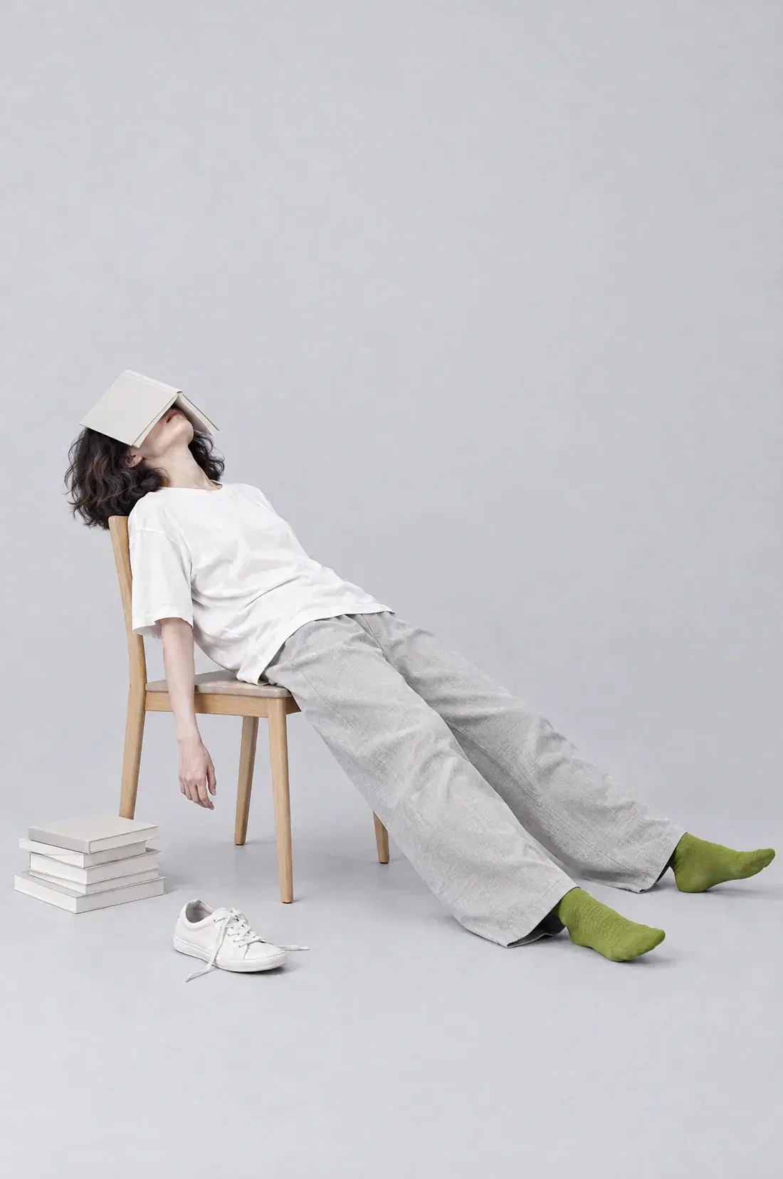 a woman slumped on a wooden chair with an open book over her face, one arm hanging down, legs stretched out in lime socks