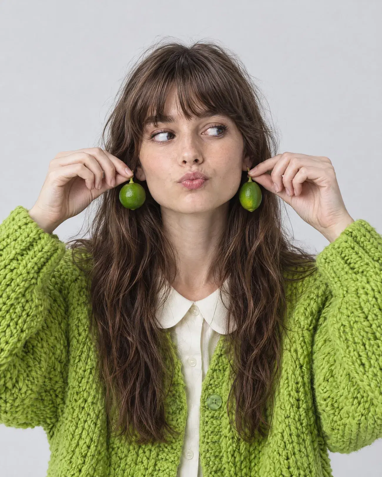 a woman in a lime knit cardigan holding two limes up like earrings
